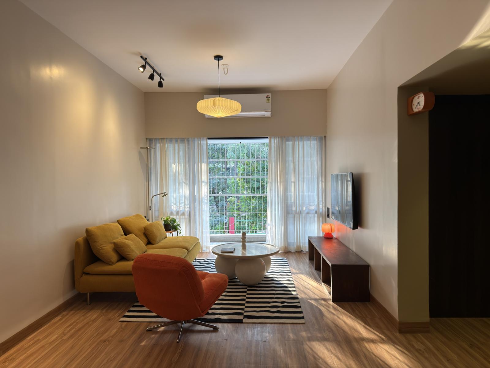 The living room: a mustard corduroy sofa, an orange armchair and a round white table on a black and white rug, with sheer curtains and trees outside.
